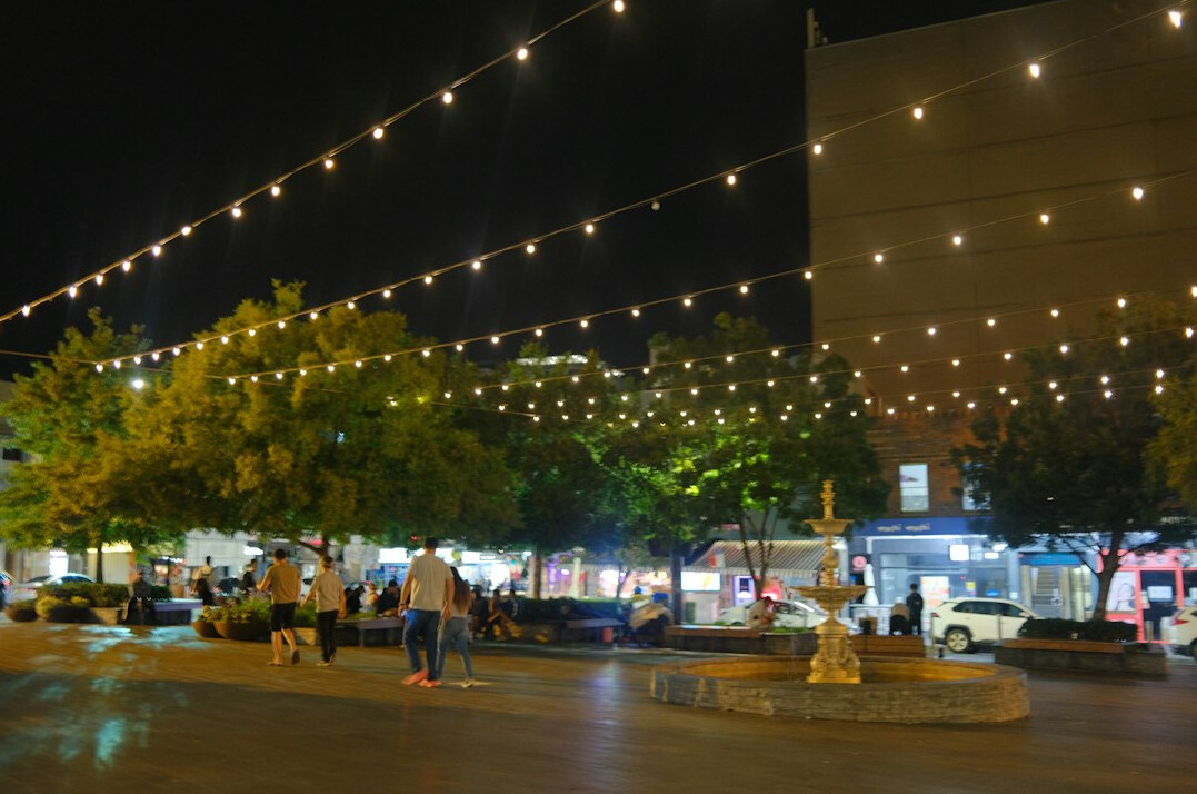 People gathered in a public plaza at dusk with string lights